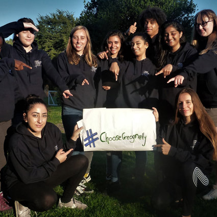 A group of students are standing holding a sign that reads 'Choose Geography'. They are pointing at it and smiling