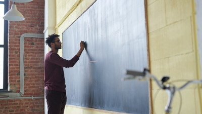 A teacher stands whilst writing on a chalk board. The rest of the room is not in view.