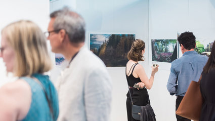People viewing a photographic exhibition displayed on white panels in a modern pavilion. The photos show forests and trees.