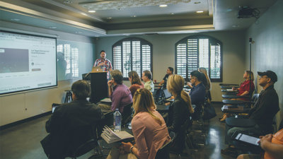 A group of people sat in a room with a projector screen with one person in the front presenting.
