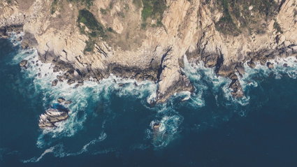 An aerial image of a rocky coastline edge, with mud and vegetation on the cliffs and waves breaking around the cliff foot.