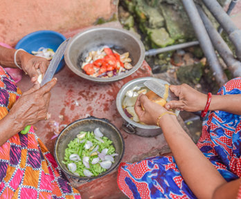 Cooking pots containing a variety of vegetables and two women cutting items