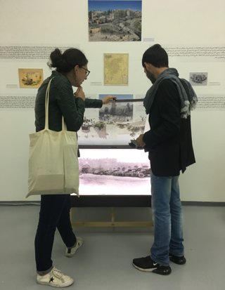 Two people stood looking at a digital image of old cars. The image is in a glass case and part of an exhibition display.