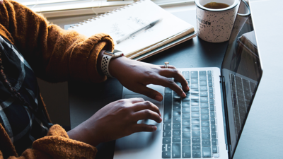 A person in a brown knitted cardigan, sitting at a table, types on a laptop. A note pad, pen and cup filled with a beverage sits next to their laptop