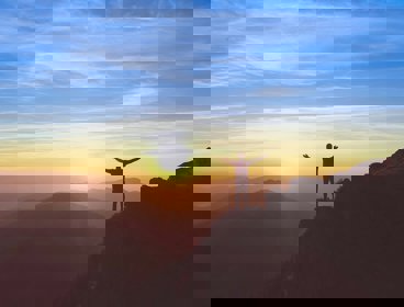 Person standing on a cliff with arms outstretched, in front of a sunset sky