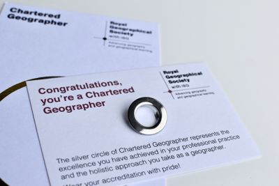 Silver circle pin awarded to Chartered Geographers shown on a white card reading 'Congratulations, you're a Chartered Geographer'