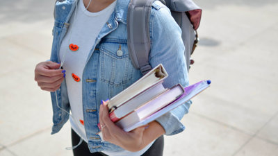 A student stands on a pavement holding two text books, a folder and notebook in one hand whilst holding on to their headphone cords with the other.
