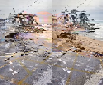A damaged house and road by the coast. Rubble lays on the beach.