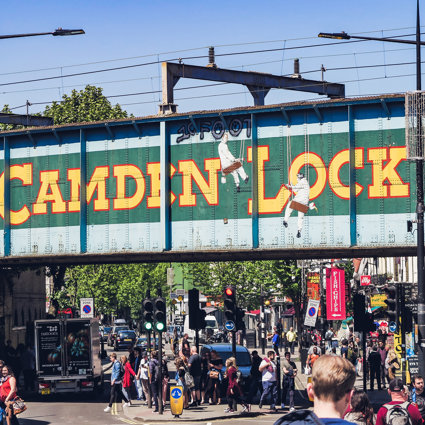 A railway bridge in Camden in London, with Camden Lock painted onto the side. There are people below the bridge crossing a busy road and heading towards local shops