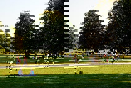 An area of parkland, showing people walking, sitting and cycling. It is a nice day and the sun is low in the sky which is casting long shadows from the trees.