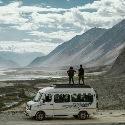 Two people are standing on top of a minibus, overlooking a dusty valley with a small river running along the bottom of it. There are mountains to the right hand side and the sky is cloudy.